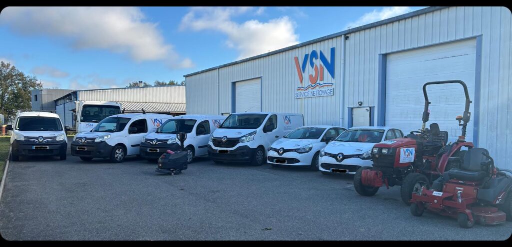VSN Service Nettoyage : la flotte de véhicules professionnelle Flotte VSN SERVICE NETTOYAGE complète : plusieurs vans, voitures et équipements (tracteur, tondeuse) devant le bâtiment de l'entreprise.